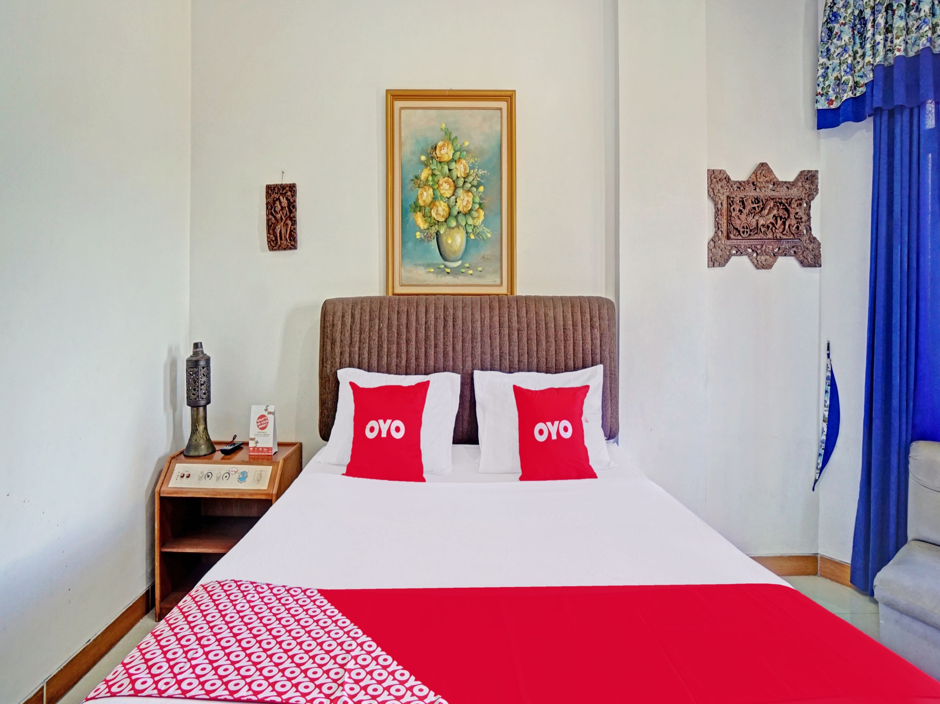 A hotel room with a bed, a nightstand, a decorative vase, a framed painting, and a blue curtain.