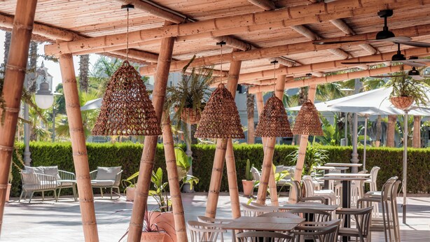Un café al aire libre con pilares de bambú, lámparas colgantes tejidas y sillas y mesas de plástico blanco.