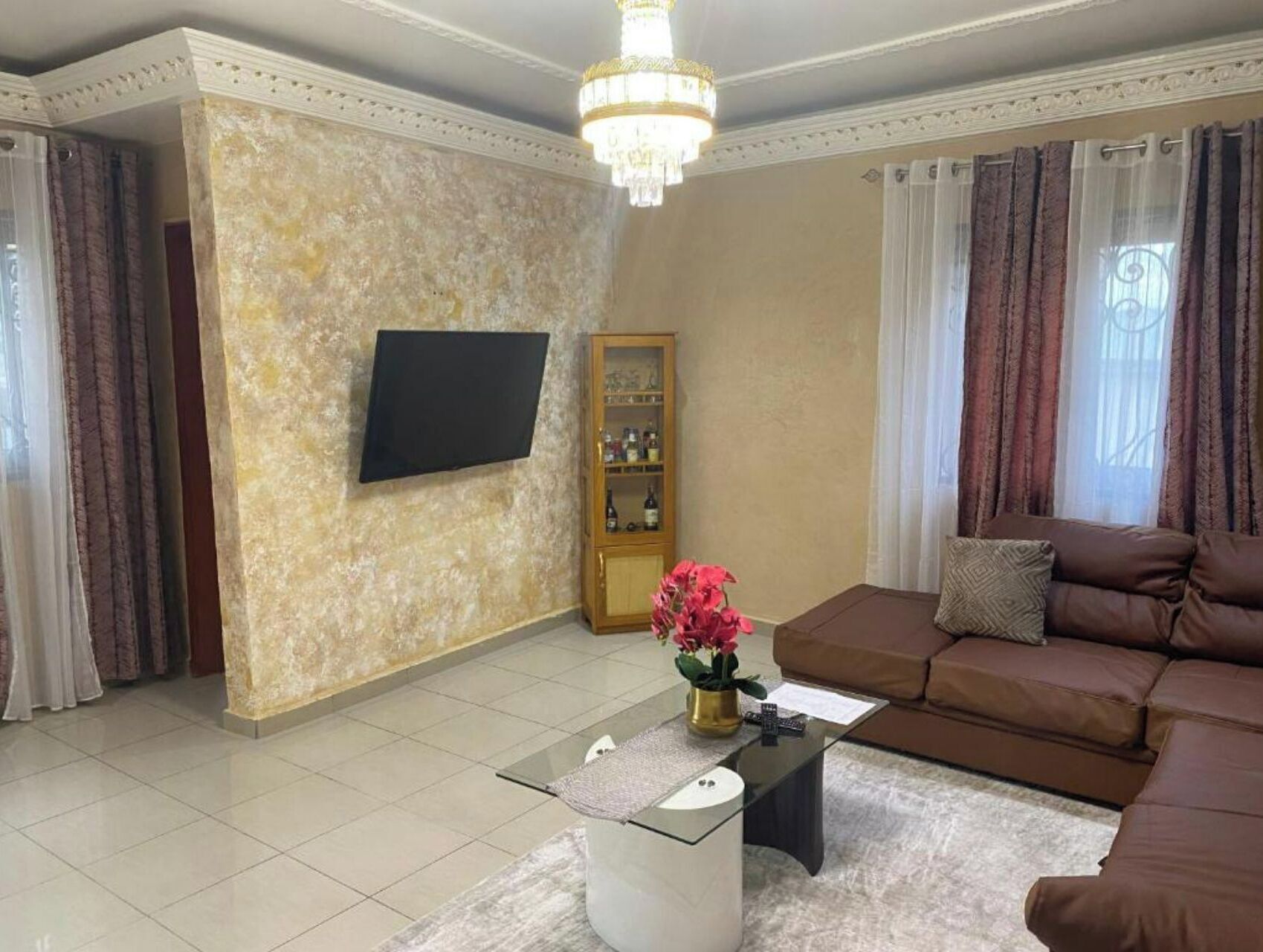 Una sala de estar con un televisor de pantalla plana montado en la pared, un sofá de cuero marrón, una mesa de centro de vidrio con un jarrón de flores y un aparador con botellas.
