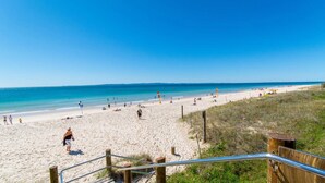 Ein Strand mit einer hölzernen Treppe, die zum Sand führt, einem klaren blauen Himmel und dem Meer im Hintergrund.