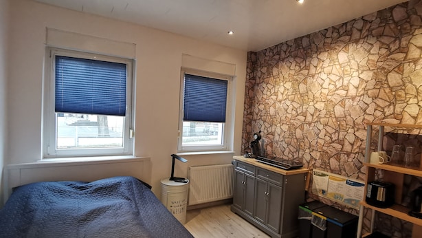 A bedroom with a bed, two windows with blinds, a stone wall, a radiator, a desk with a computer, and a shelf with mugs. - Hübsches Zimmer mit Eigenem Bad und Kochecke, Gute Verkehrsanbindung (Swisttal)