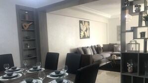 A dining area with a table set for six, a grey sofa, and a bookshelf with decorative items. - Appartement 105m2 Haut Standing Quartier du Parc Proche Plage (Mohammédia)