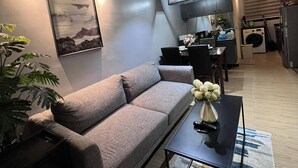 Un salon moderne avec un canapé gris, une table basse et une plante. On aperçoit à l’arrière-plan un coin cuisine avec une table à manger et des chaises.