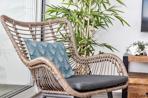 Een rieten stoel met een kussen met een geometrisch patroon, geplaatst nabij een grote bamboeplant in een kamer met een raam en een houten bijzettafeltje met decoratieve objecten.
