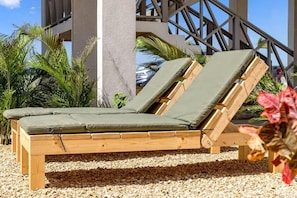 A wooden lounge chair with a green cushion, positioned on a gravel surface with a palm tree and a building in the background.