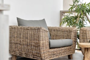 A wicker chair with grey cushions, a wooden table, and a potted plant in a room with a white wall and a wooden railing.