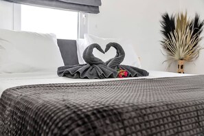 A neatly made bed with a heart-shaped towel arrangement, a decorative plant, and a window with a view.