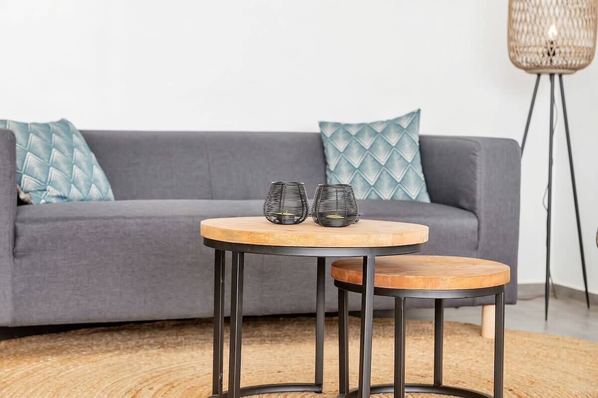A round wooden table with metal legs in front of a grey sofa with patterned cushions.