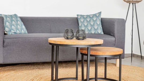 Une table ronde en bois, dotée de pieds en métal, se trouve devant un canapé gris agrémenté de coussins à motifs.