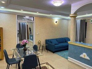 Una sala de estar con un sofá azul, una mesa de comedor de vidrio con sillas y un jarrón decorativo con flores.