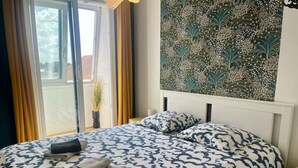 A bedroom with a bed, a window with curtains, a wall with a floral pattern, and a plant. - Appt Centre de Courseulles - 100m de la Plage (Courseulles-sur-Mer)