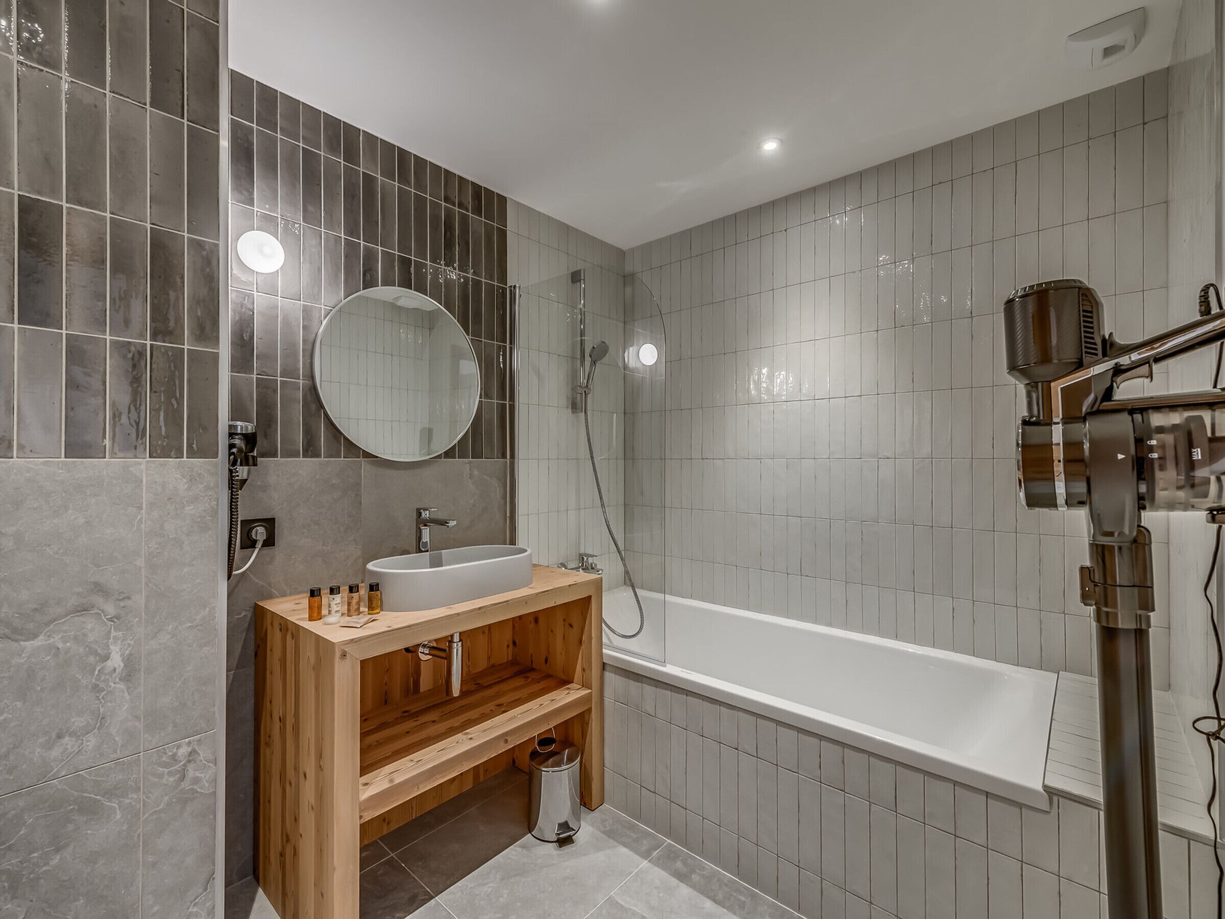 Un baño moderno con tocador de madera, espejo redondo, bañera y zona de ducha.