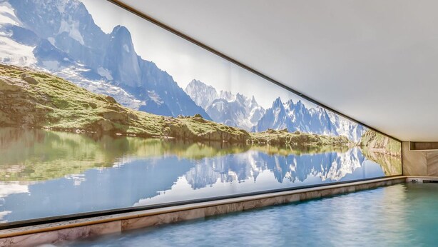 A serene indoor swimming pool with a panoramic view of a mountain lake and peaks.