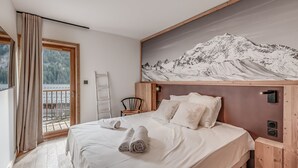 Ein Schlafzimmer mit Bett, Holz-Kopfteil, Wandbild mit Bergmotiv, Fenster mit Vorhängen und Blick ins Freie.