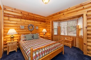 A log cabin bedroom with a plaid bedspread, two bedside lamps, a wooden dresser, and a window with blinds.