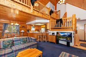 Interior de una cabaña con escalera de madera, un sofá, una televisión y una zona de cocina.