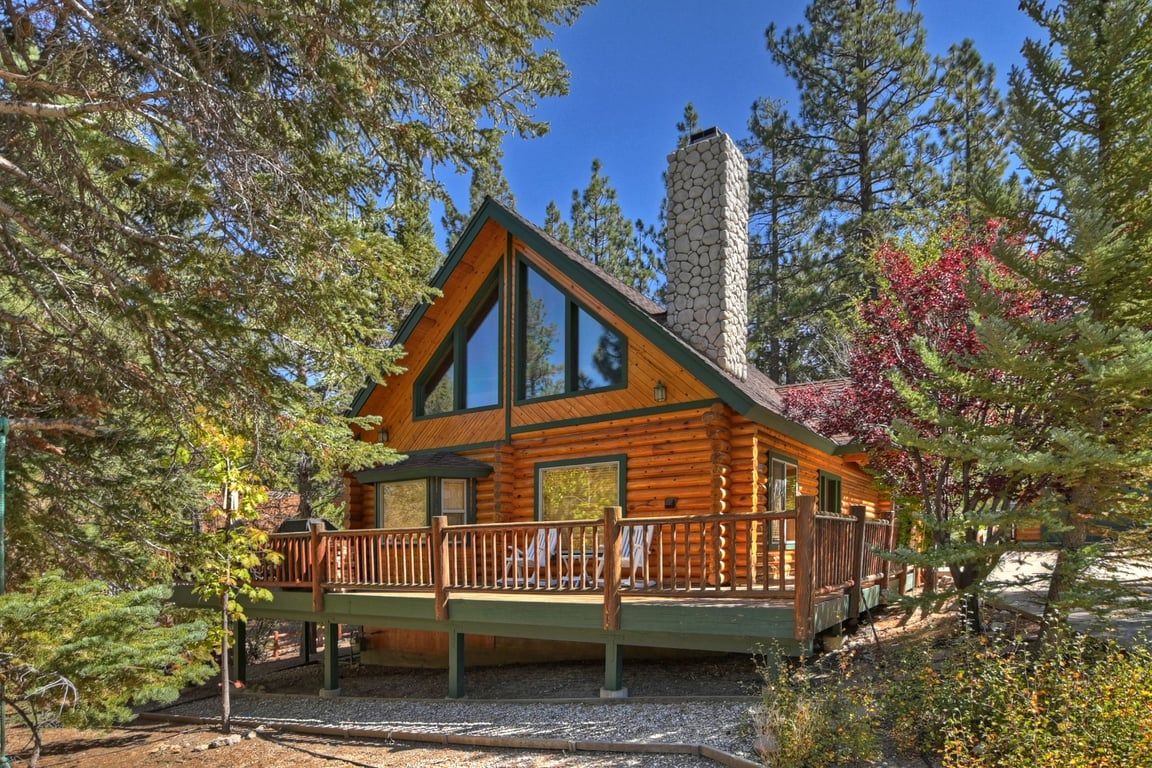 A log cabin with a stone chimney, a deck with railings, and surrounded by trees.