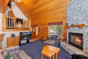 A log cabin interior with a stone fireplace, plaid furniture, a television, and a wooden staircase.