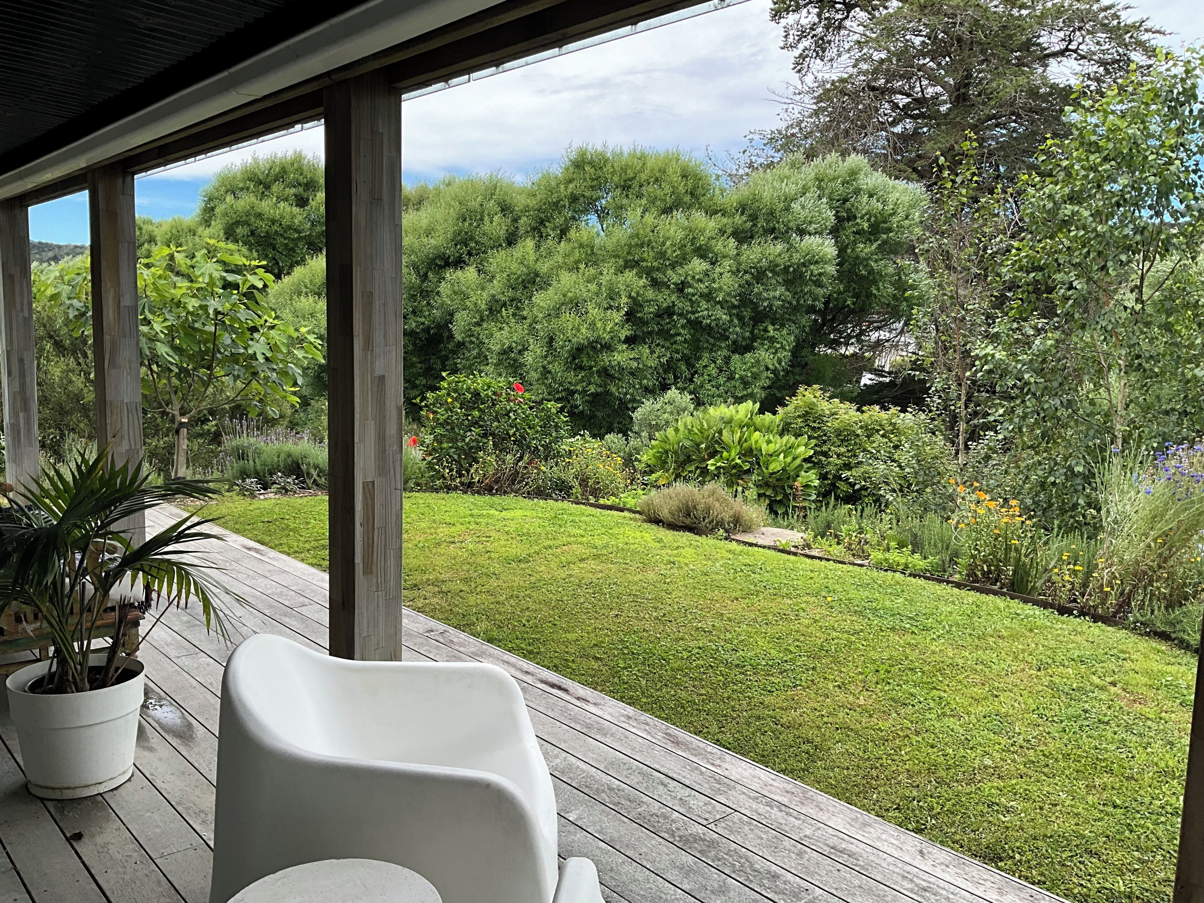 A covered outdoor area with a wooden deck, potted plants, and a view of a well-maintained garden with various shrubs and trees.