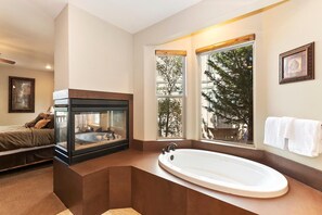 Een moderne badkamer met een vrijstaand bad, een glazen haard en grote ramen met uitzicht op bomen.