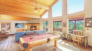 A spacious living area with a pool table, a stone fireplace, and large windows offering views of the outdoors.