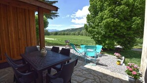 An outdoor patio with a stone table and chairs, a wooden pergola, and a view of a green field and mountains.
