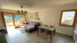A spacious living area with a sofa, a dining table with chairs, a balcony with a view of greenery, and a ceiling light fixture.