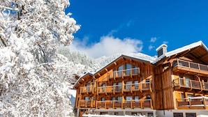 Eine Holzhütte mit mehreren Balkonen, umgeben von schneebedeckten Bäumen und wolkenlosem, blauem Himmel.