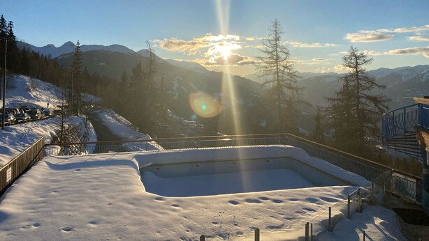 Zona exterior cubierta de nieve con piscina, rodeada de montañas y árboles.