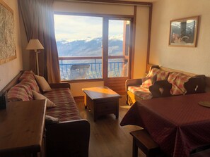 A cozy room with a wooden floor, a sofa, a table, and a window with a view of snow-capped mountains.