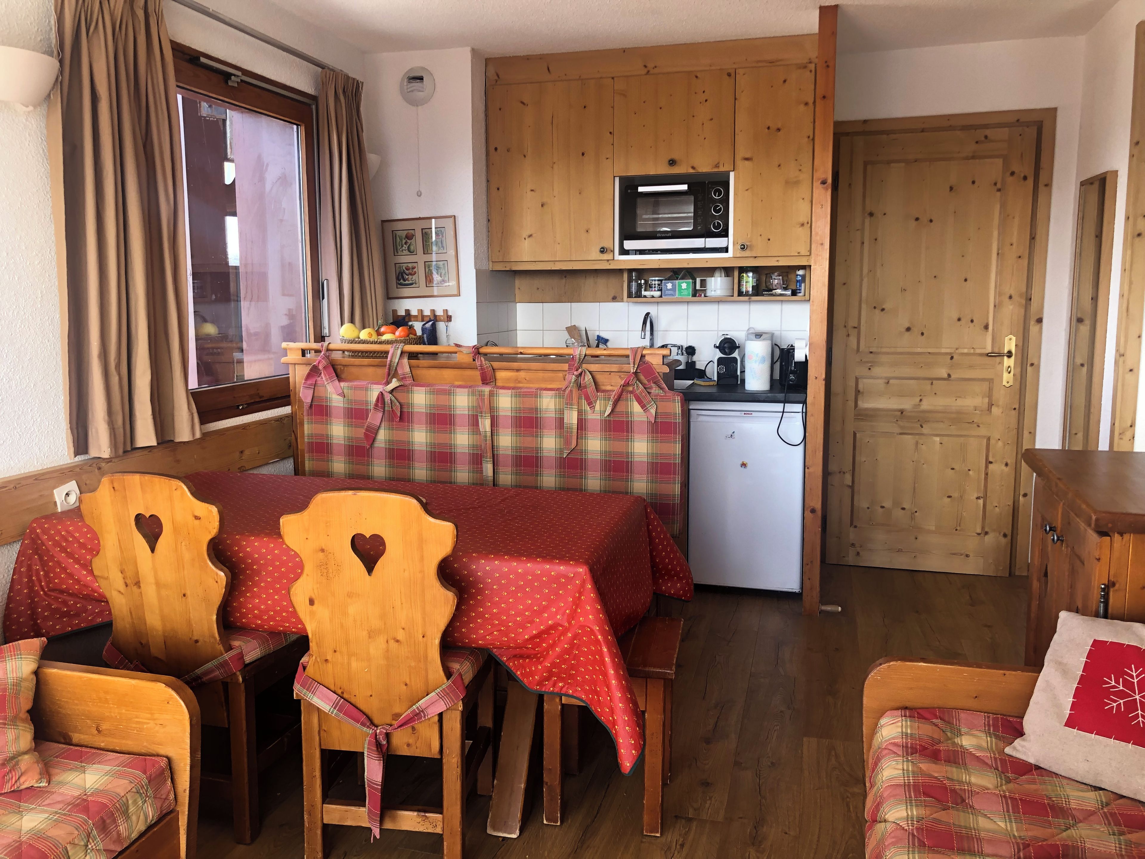 A compact kitchen with a microwave, refrigerator, and wooden cabinets. There is a dining area with a table and chairs, and a window with curtains.