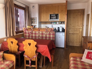 Eine kompakte Küche mit Mikrowelle, Kühlschrank und Holzschränken. Es gibt eine Essecke mit Tisch und Stühlen sowie ein Fenster mit Vorhängen.