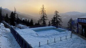 Zona exterior cubierta de nieve con una piscina, rodeada de montañas y árboles.