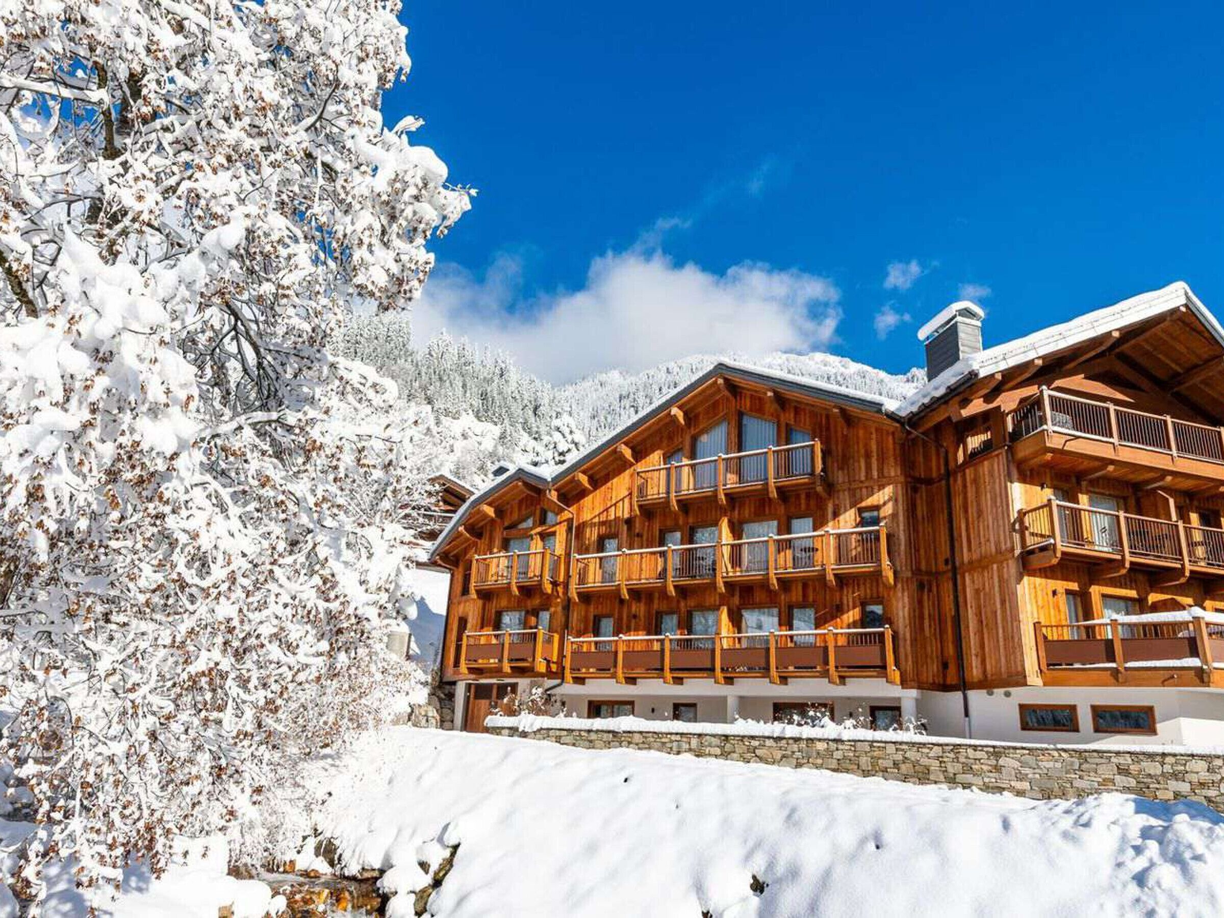Eine Holzhütte mit mehreren Balkonen, umgeben von schneebedeckten Bäumen und wolkenlosem, blauem Himmel.