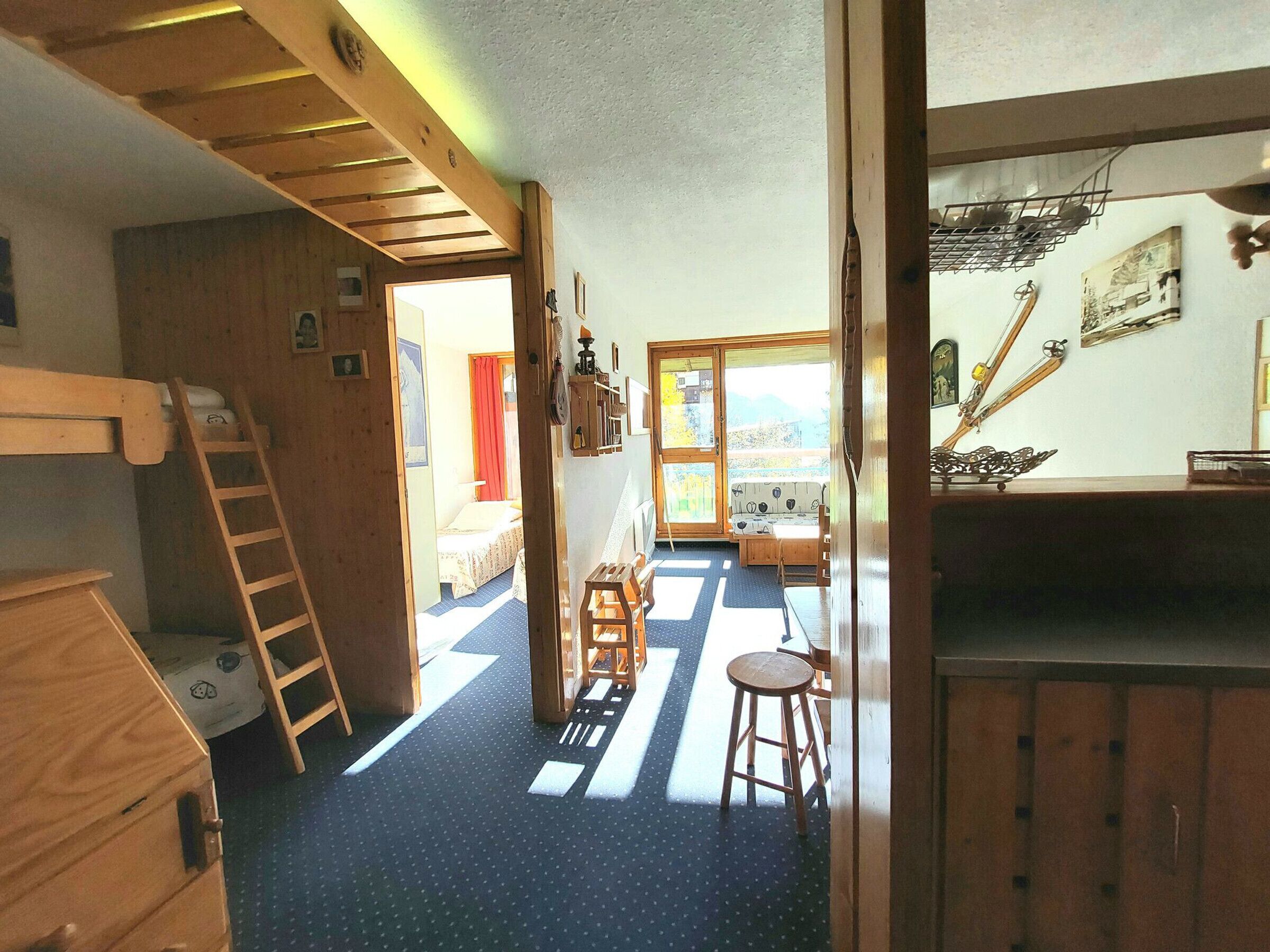 Een kamer met een stapelbed, een ladder, een klein tafeltje met een krukje, een raam met uitzicht en een deur die naar een andere kamer leidt.