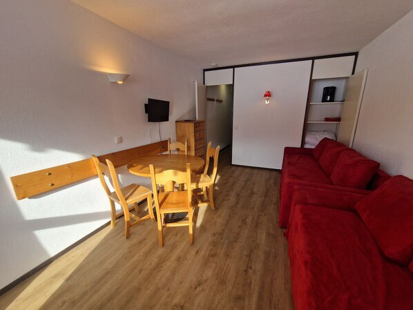 Un espace de vie compact comprenant une table à manger, des chaises, un canapé rouge, un téléviseur et un coin cuisine.