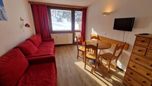 A room with a red sofa, wooden dining table, chairs, a TV, and a window with a view of a snowy landscape. - Bright studio with balcony, ski-in/ski-out, sleeps 4 (La Plagne)