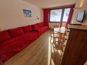 Habitación con sofá rojo, mesa de comedor de madera, sillas, televisor y ventana con vistas a un paisaje nevado.