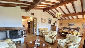 A spacious living room with wooden beams, floral-patterned sofas, a wooden coffee table, and a dining area with a table set for four.