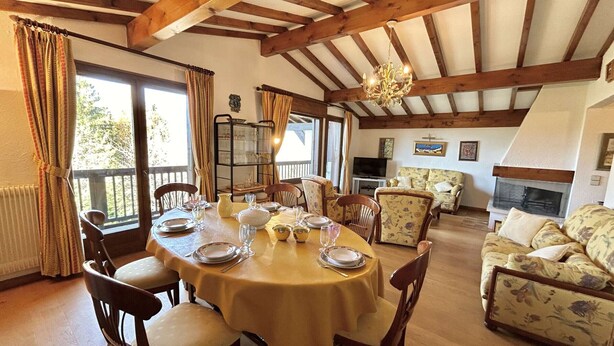 A dining area with a round table set for six, surrounded by wooden chairs. There is a sofa and a fireplace in the background.