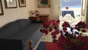 Un saló amb un sofà, una taula amb un ram de flors vermelles i una porta que dona a un balcó amb vista al mar.