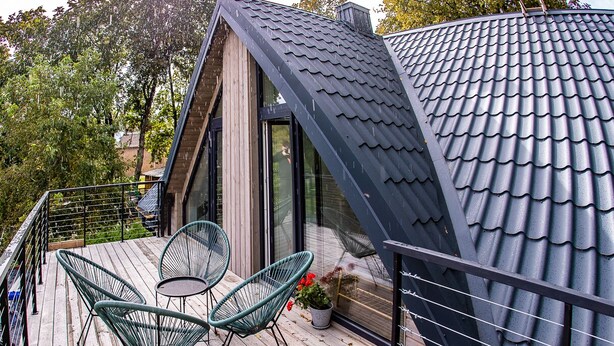 Una casa moderna con techo inclinado, una terraza de madera y dos sillas de estructura metálica verde.
