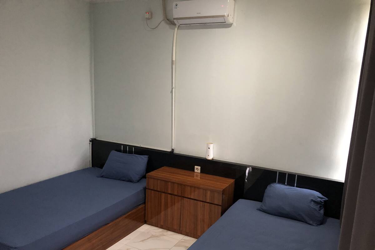 Una habitación pequeña y sencilla con dos camas individuales, una mesita de noche de madera y un aire acondicionado instalado en la pared.