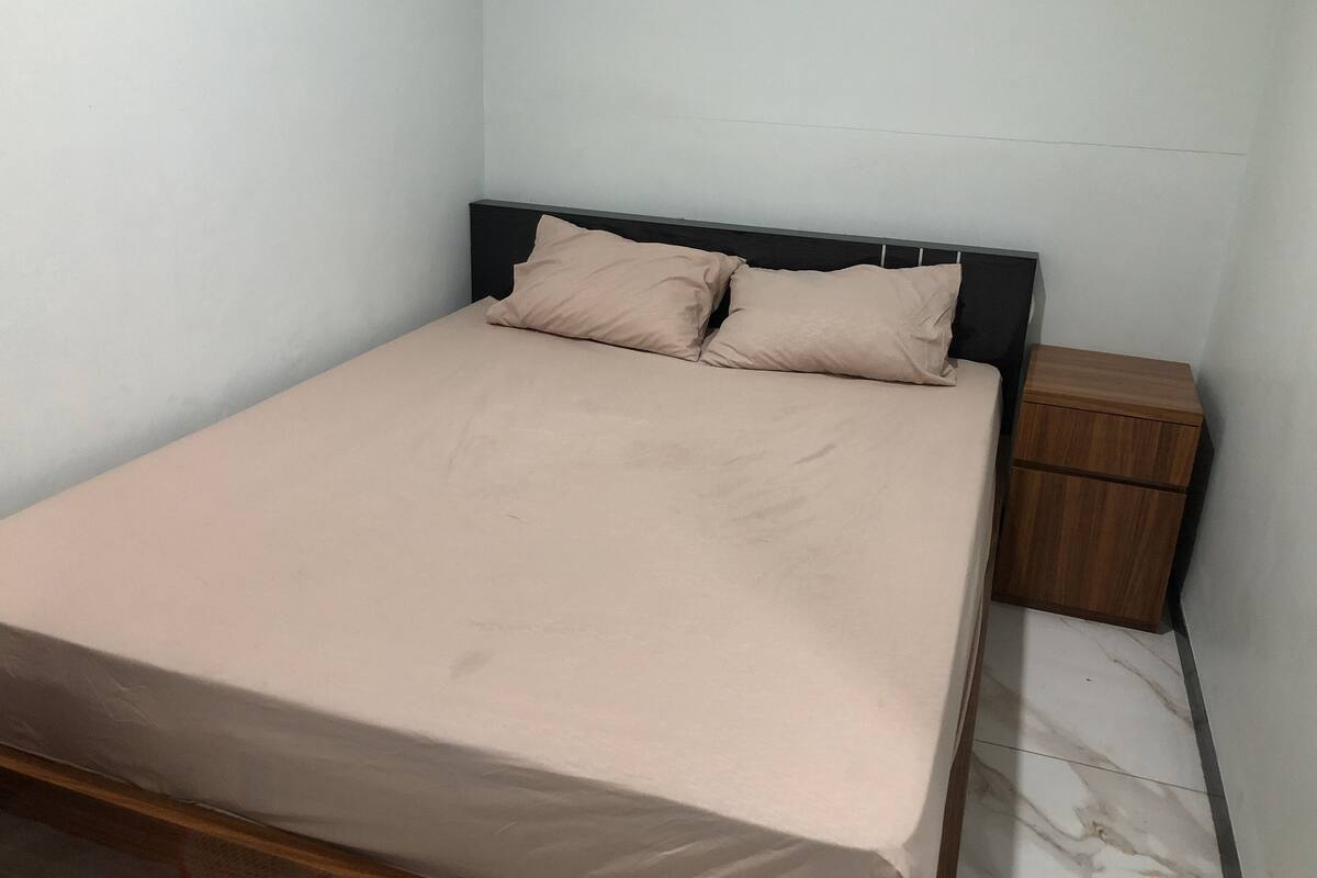 Una cama individual con colchón color beige y dos almohadas, una mesita de noche de madera a su lado, y suelo de mármol.