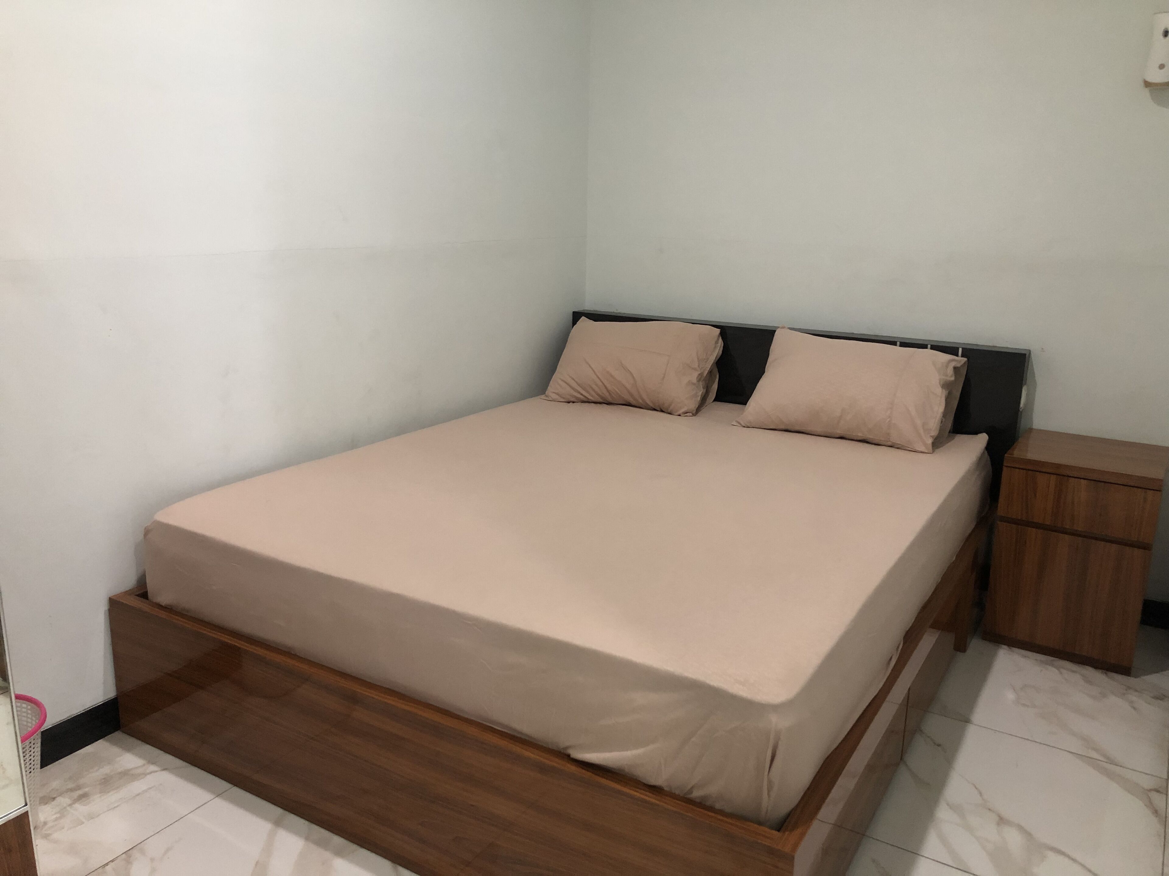 A neatly made bed with a wooden frame and a beige mattress, a small wooden nightstand beside it, and a white tiled floor.
