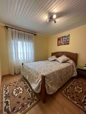 A bedroom with a bed, two rugs, a nightstand, a lamp, a painting, and a window with curtains. - Casa Violeta (Águeda)