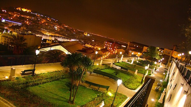 Un espace jardin bien éclairé, agrémenté de sentiers, de bancs et de statues, entouré de bâtiments et offrant une vue sur le paysage urbain la nuit.