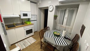 Une cuisine compacte avec des meubles blancs, un four à micro-ondes, un petit four et un évier. On y trouve une table à manger avec une nappe à rayures et des chaises. Une horloge est accrochée au mur, et il y a une fenêtre avec des rideaux.