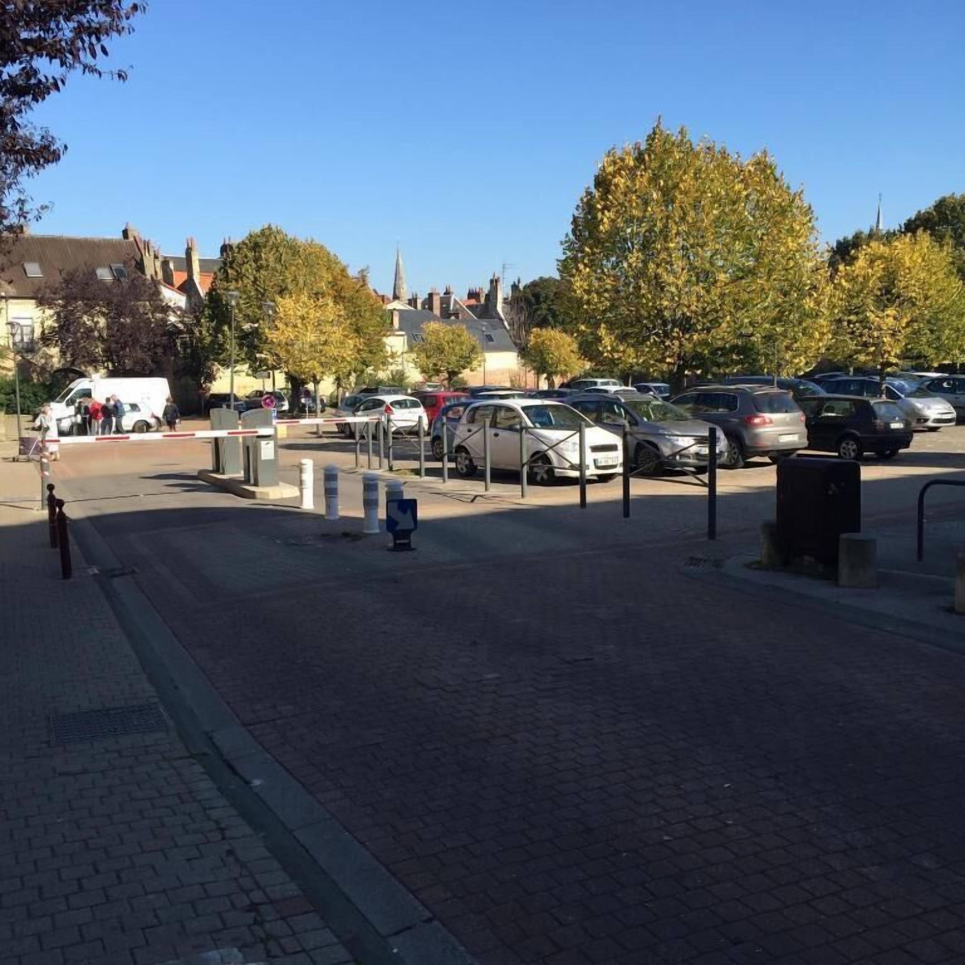 Un espace de stationnement avec des voitures et un fourgon blanc, entouré d’arbres et d’immeubles.