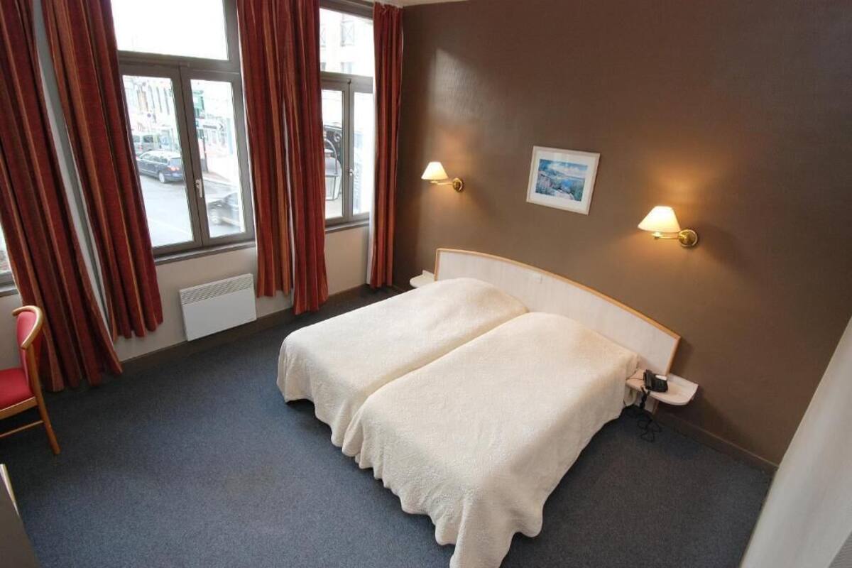 A hotel room with a double bed, two bedside lamps, a window with curtains, a chair, and a radiator.